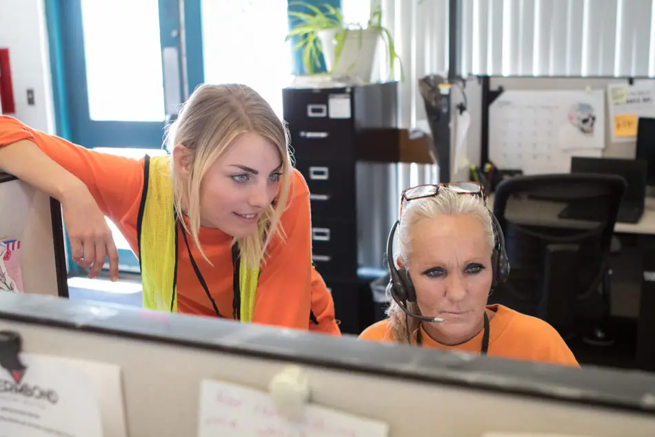 Two workers at a call center workstation, one wearing a headset