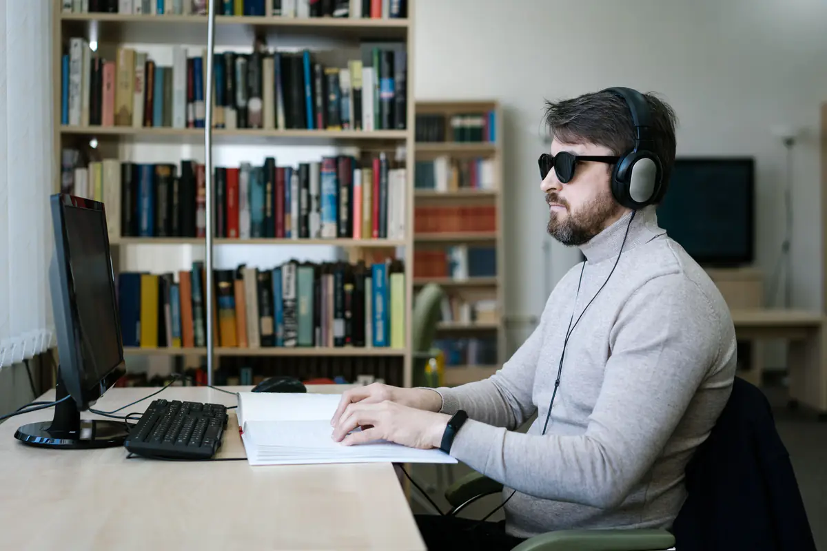 A man wearing headphones working with a braille book and a computer in a library