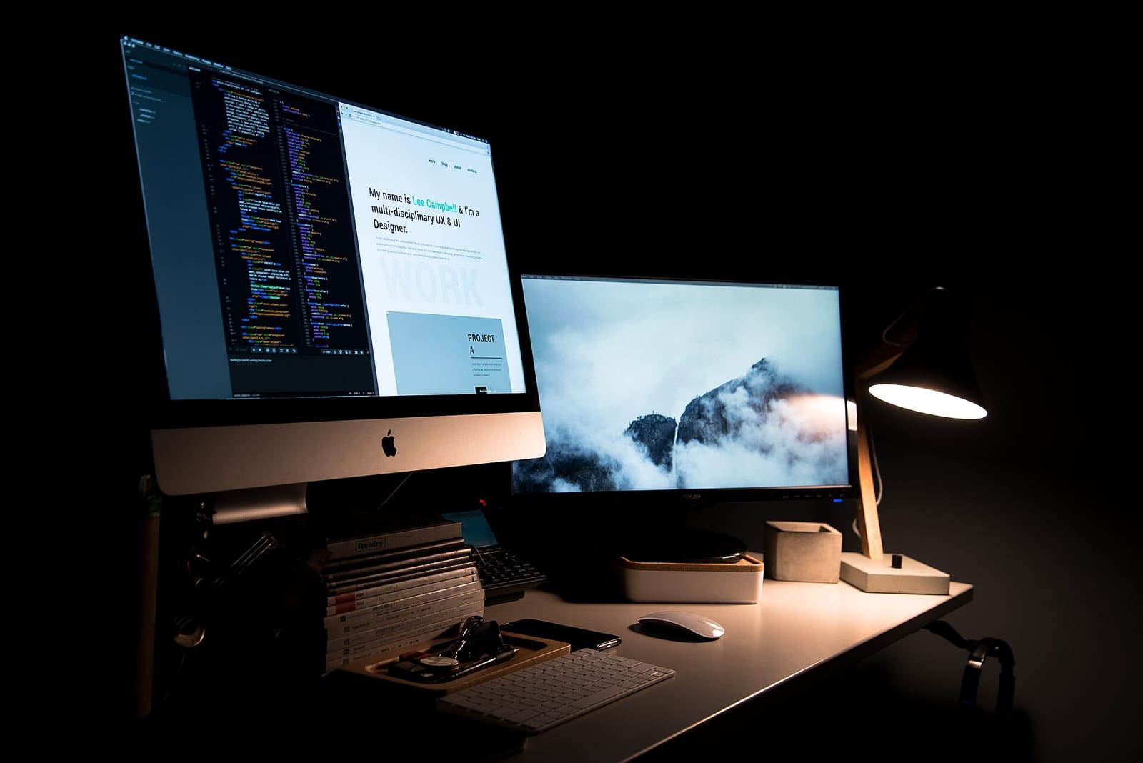 A designer's desk with two monitors, keyboard and lamp in a dark room