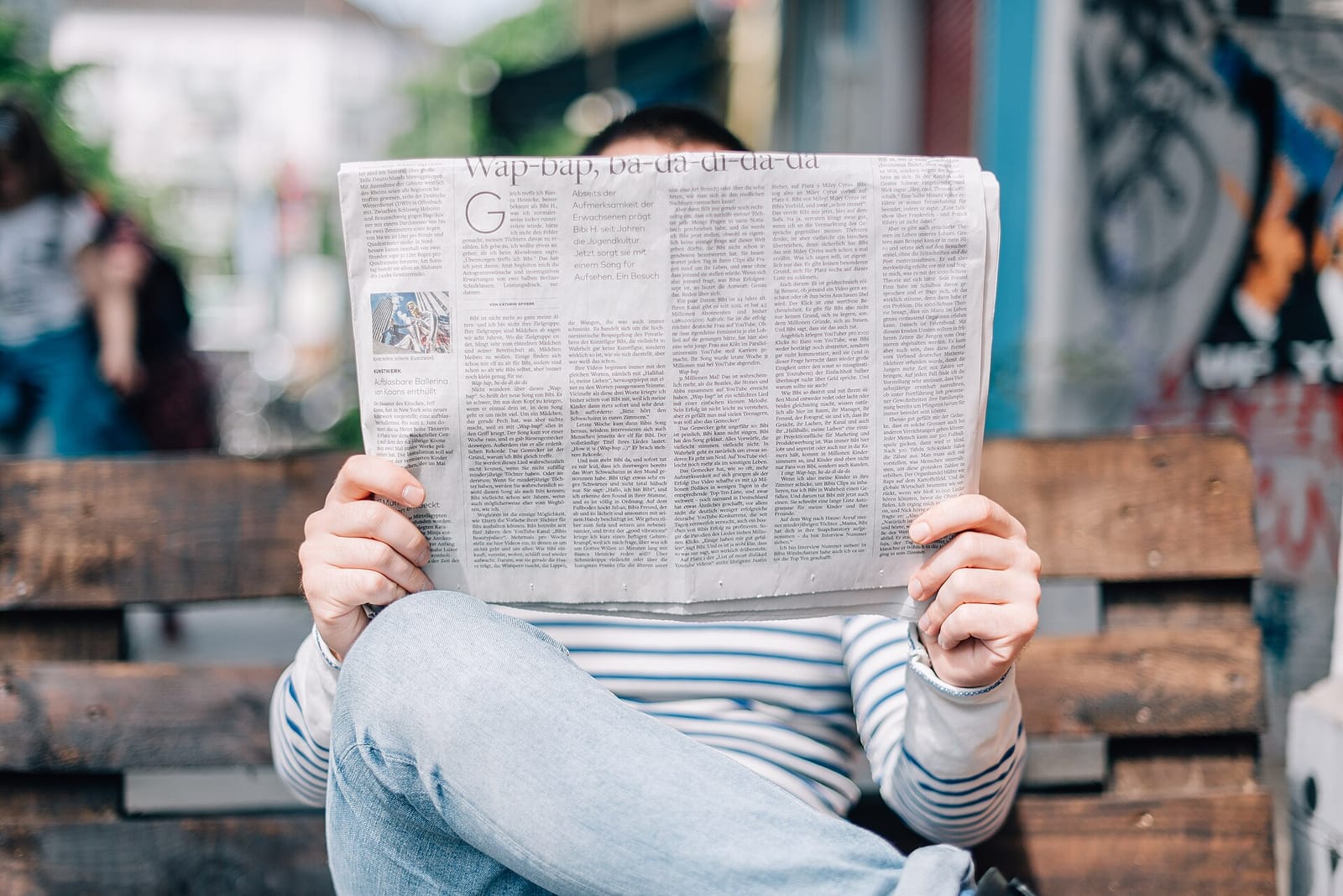 A person sitting on a wooden bench reading an open newspaper