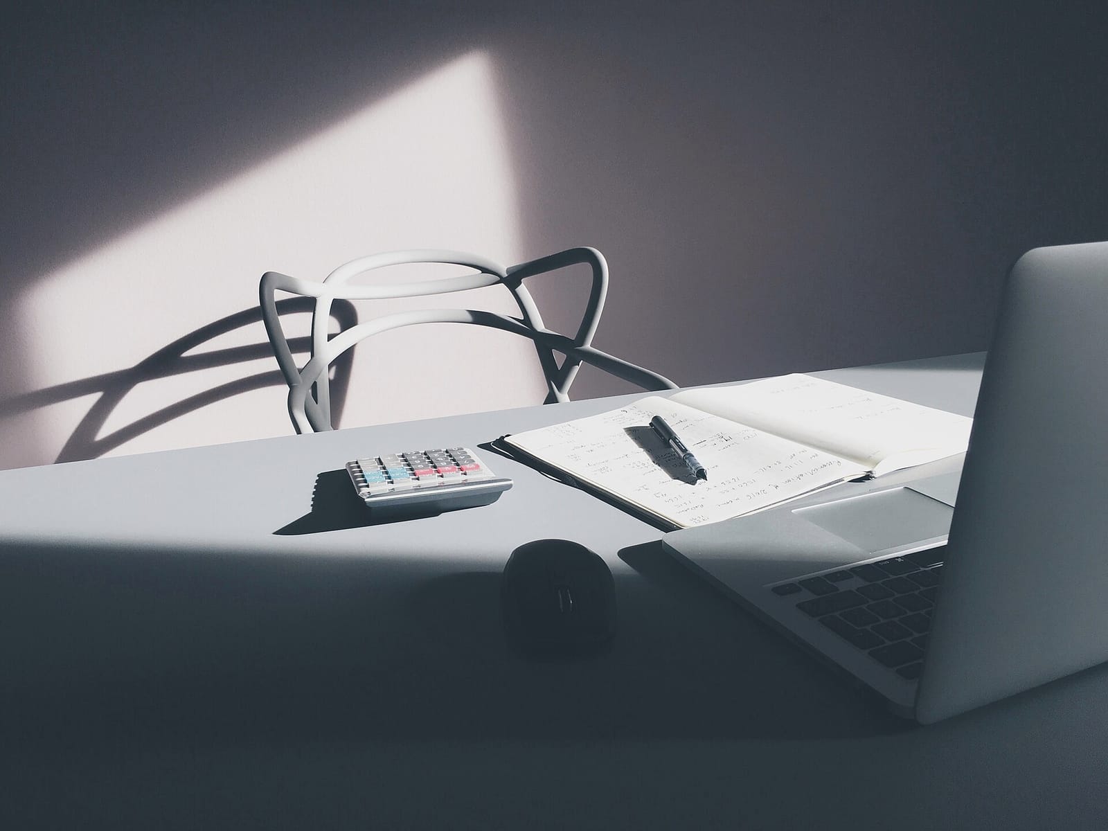A desk with a calculator, an open notebook and a laptop in a sunlit room