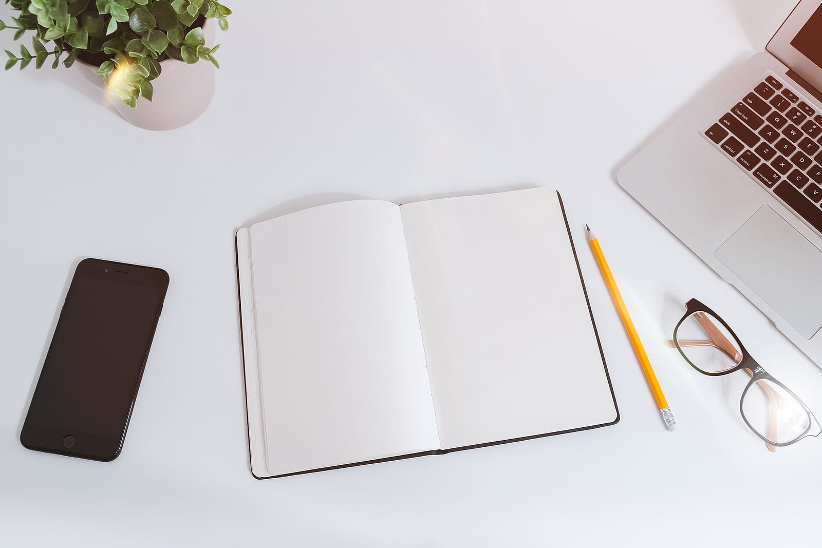 A white desk with an open notebook, pencil, glasses, smartphone and laptop