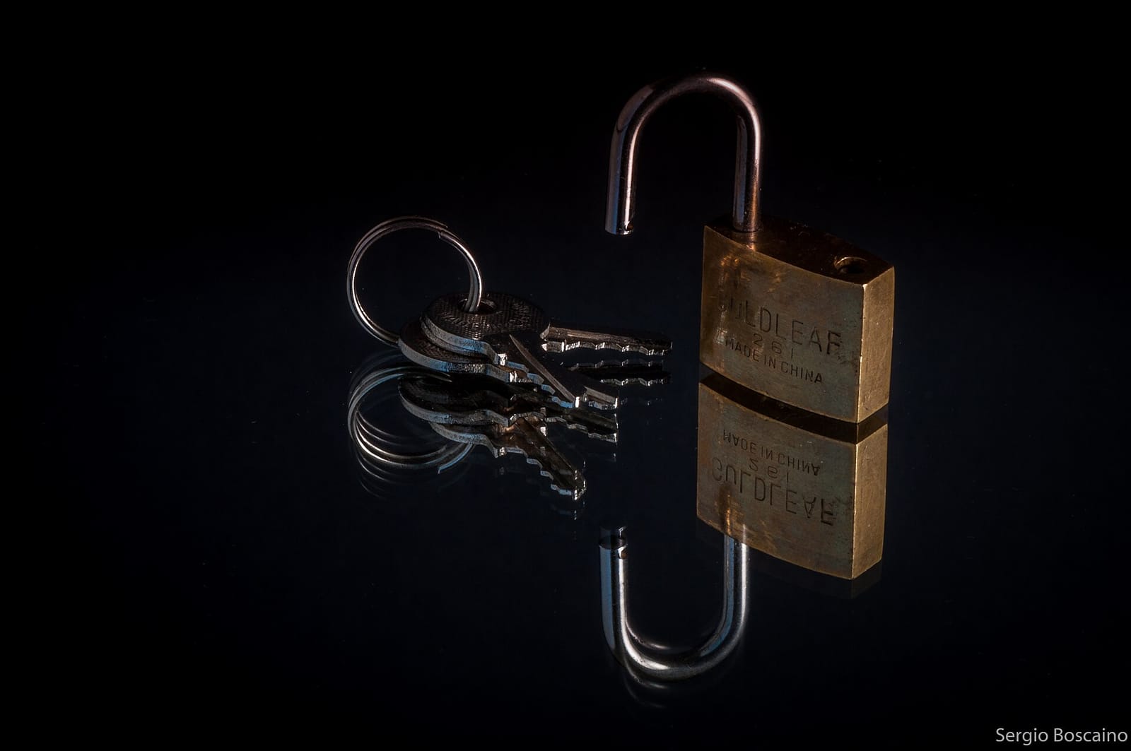 An open brass padlock next to a set of keys on a black reflective surface