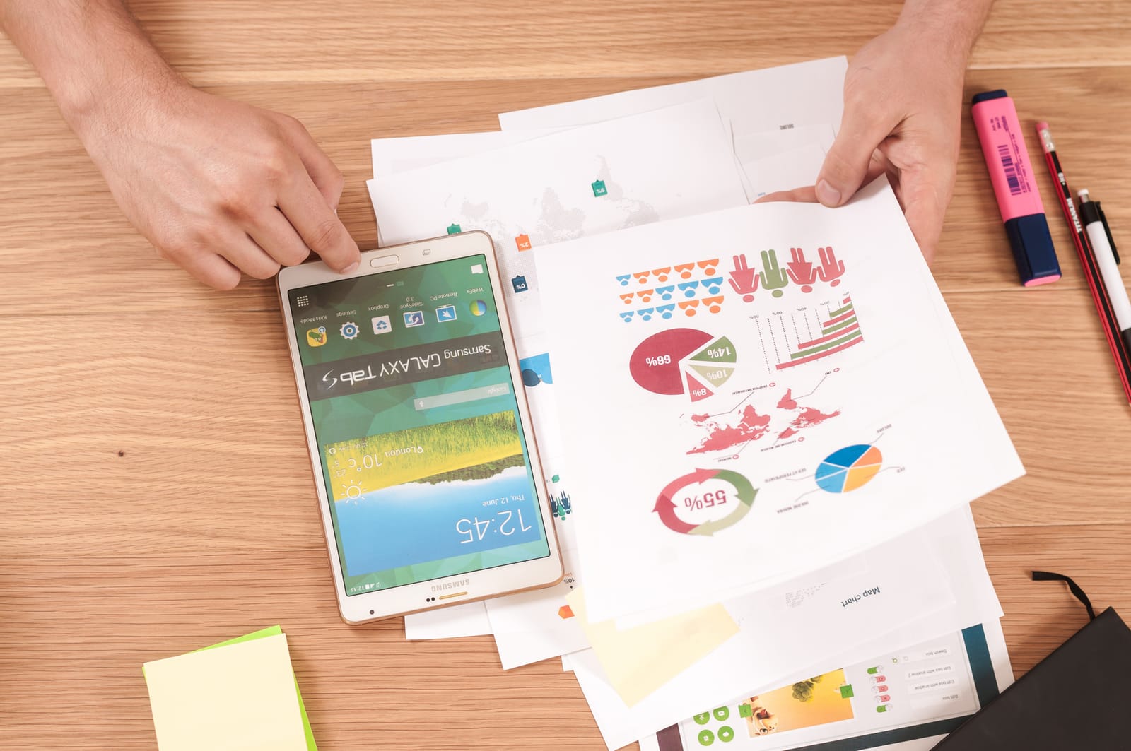 Hands holding a tablet over printed charts and graphs on a wooden desk