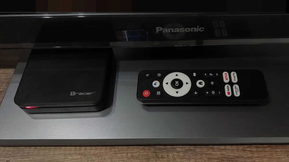 A small black Android TV set-top box and its remote control placed in front of a television