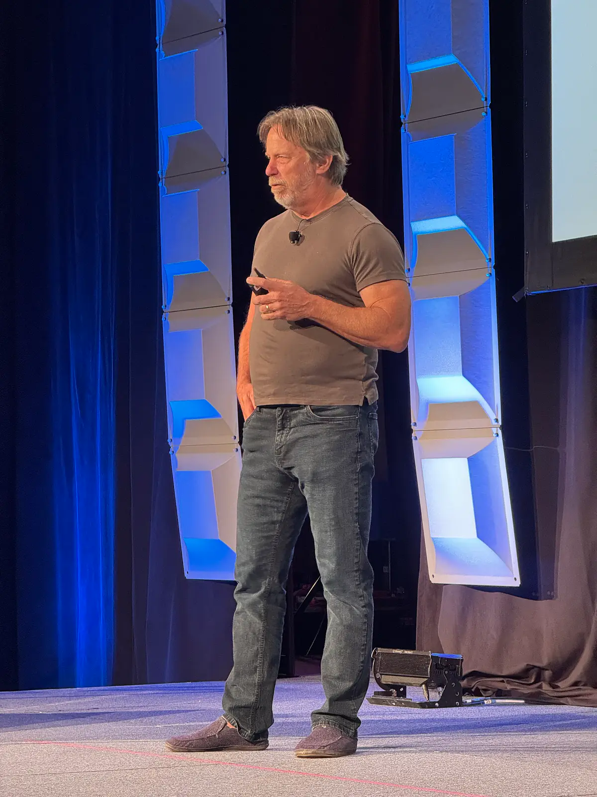 Jim Keller speaking on a stage with a microphone headset in front of blue lit panels